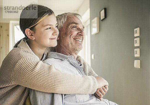 Lächelndes Mädchen umarmt Großvater zu Hause von hinten