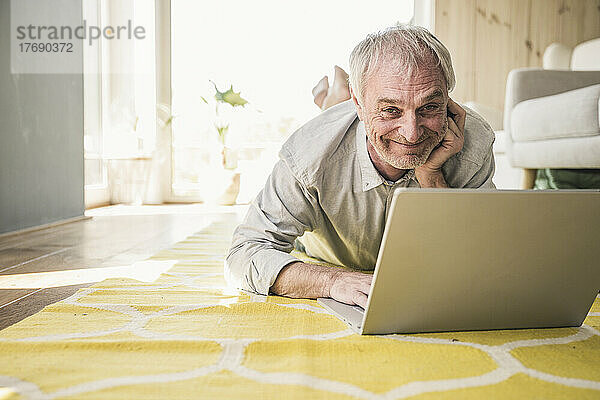 Glücklicher älterer Mann mit Laptop  der zu Hause auf dem Teppich liegt