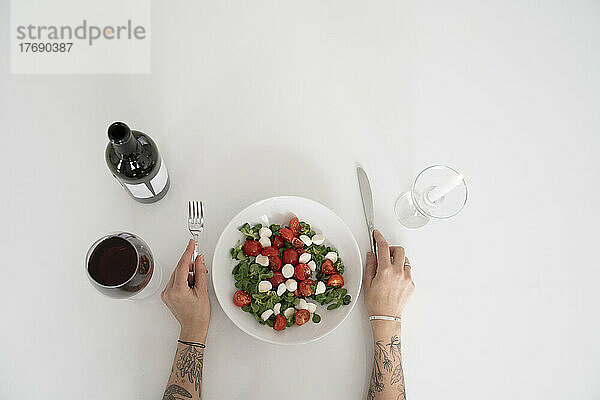 Hände einer Frau  die Besteck neben einer Salatschüssel und Wein auf einem weißen Tisch hält