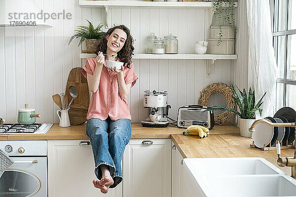 Glückliche Frau beim Frühstück  die zu Hause auf der Küchentheke sitzt