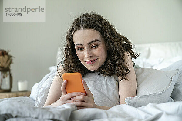 Lächelnde junge Frau  die zu Hause im Bett ihr Mobiltelefon benutzt