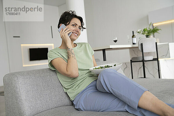 Lächelnde Frau hält Salatschüssel in der Hand und telefoniert zu Hause