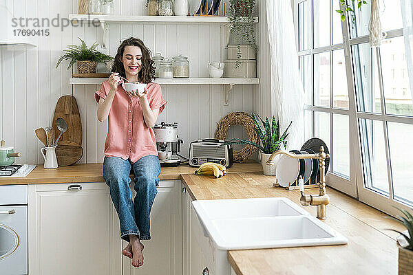 Glückliche junge Frau frühstückt zu Hause auf der Küchentheke