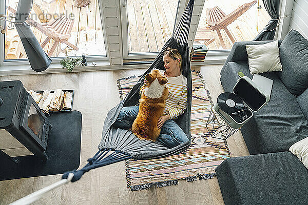 Frau mit Hund liegt zu Hause auf Hängematte im Wohnzimmer