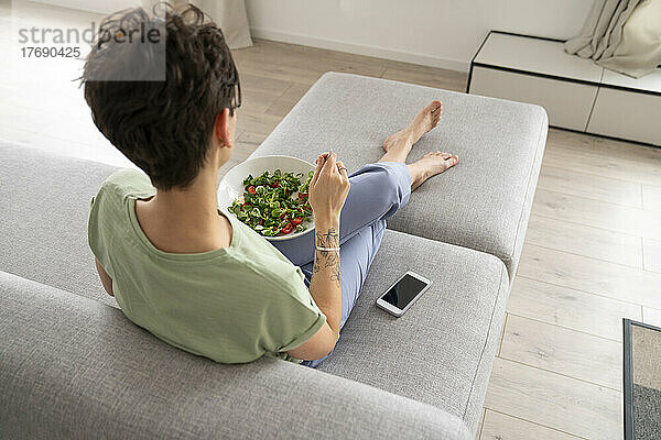 Frau isst Salat und sitzt zu Hause mit dem Handy auf dem Sofa