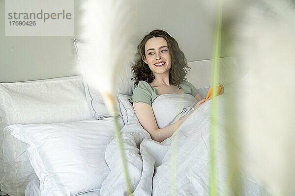 Glückliche Frau mit Smartphone sitzt zu Hause im Bett