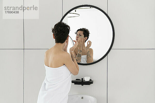 Frau in Handtuch gehüllt  Augenmaske auftragend und in den Spiegel schauend