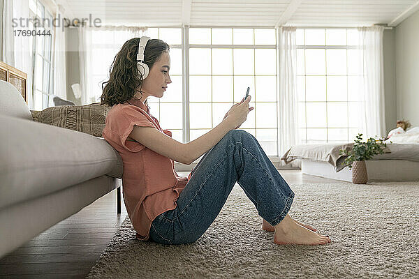 Junge Frau sitzt zu Hause auf dem Teppich und nutzt ihr Smartphone