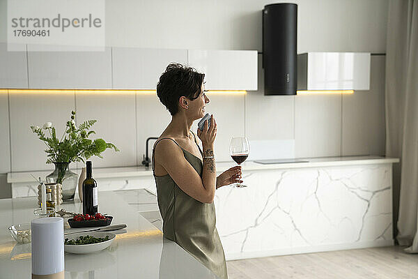 Glückliche Frau hält Weinglas in der Hand und spricht über den Handy-Lautsprecher in der Küche