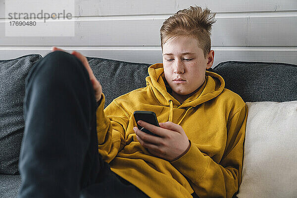 Junge im gelben Kapuzenshirt benutzt Smartphone auf Sofa zu Hause