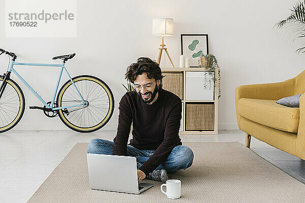 Glücklicher Mann mit Laptop  der zu Hause auf dem Teppich sitzt