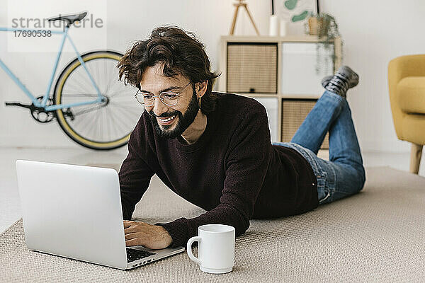 Lächelnder Mann mit Laptop  der zu Hause neben einer Kaffeetasse auf dem Teppich liegt