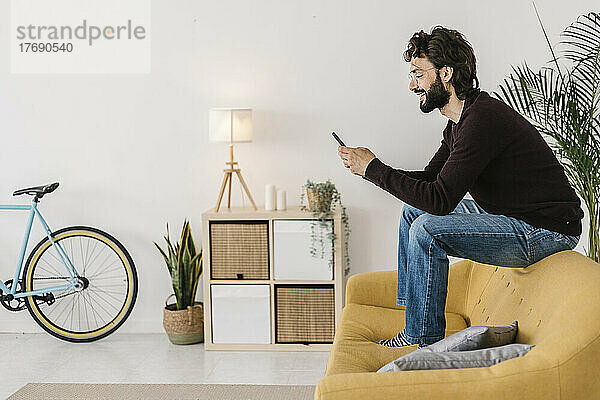 Glücklicher Mann mit Bart  der zu Hause auf dem Sofa im Wohnzimmer sitzt und sein Mobiltelefon benutzt