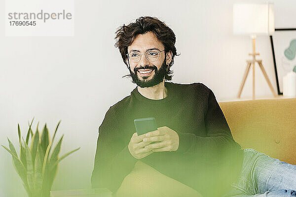 Lächelnder Mann mit Mobiltelefon sitzt zu Hause auf dem Sofa