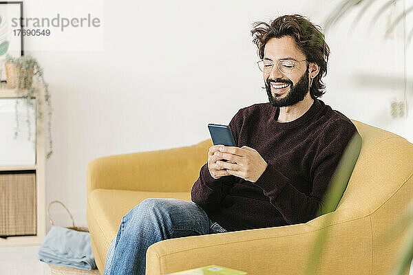 Glücklicher Mann mit Brille und Smartphone  der zu Hause auf dem Sofa sitzt