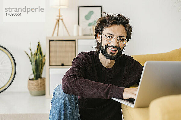 Lächelnder Mann mit Laptop sitzt zu Hause am Sofa