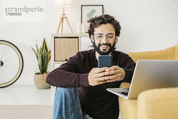 Lächelnder Mann  der zu Hause auf dem Sofa sitzt und sein Mobiltelefon benutzt