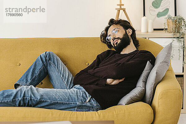 Nachdenklicher bärtiger Mann mit verschränkten Armen entspannt sich zu Hause auf dem Sofa