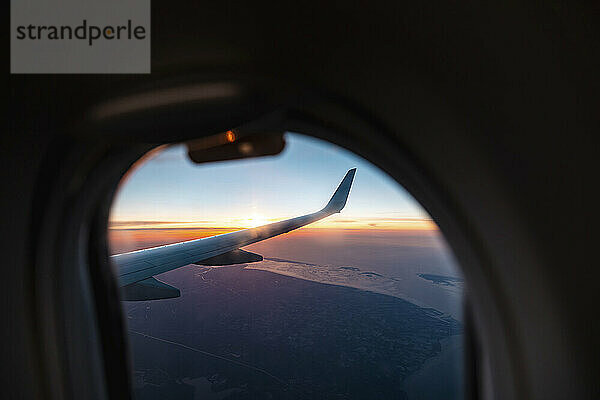 Wing of airplane flying against setting sun seen through porthole