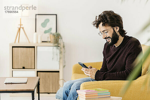 Lächelnder Mann mit Brille und Smartphone sitzt zu Hause auf dem Sofa