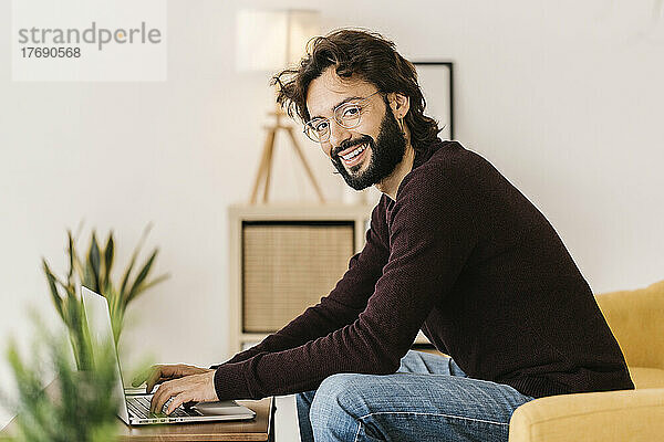 Lächelnder bärtiger Hipster-Mann mit Laptop sitzt zu Hause auf dem Sofa