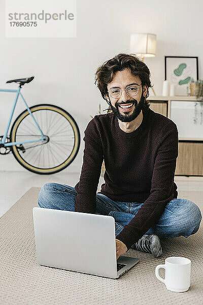 Glücklicher Mann  der zu Hause mit Laptop und Kaffeetasse auf dem Teppich sitzt