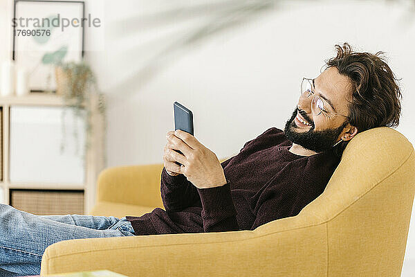 Glücklicher Mann  der zu Hause auf dem Sofa sitzt und sein Smartphone benutzt