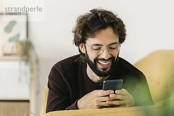 Lächelnder bärtiger Mann  der zu Hause auf dem Sofa sein Mobiltelefon benutzt