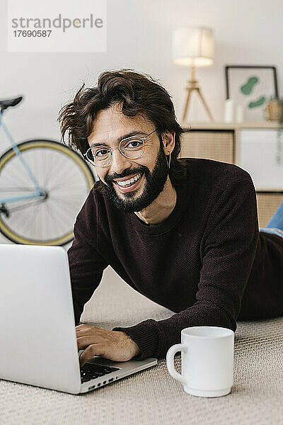 Lächelnder Mann mit Laptop und Kaffeetasse  der zu Hause auf dem Teppich liegt