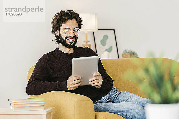 Glücklicher Mann bei Videoanruf über Tablet-PC  der zu Hause im Wohnzimmer sitzt