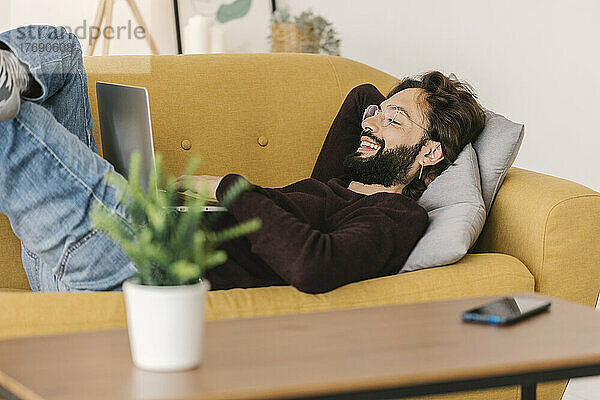 Lächelnder Mann mit Laptop  der zu Hause auf dem Sofa im Wohnzimmer liegt