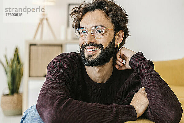 Glücklicher Mann mit Bart sitzt zu Hause am Sofa im Wohnzimmer