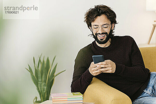 Glücklicher Mann mit Bart  der zu Hause auf dem Sofa sitzt und sein Mobiltelefon benutzt