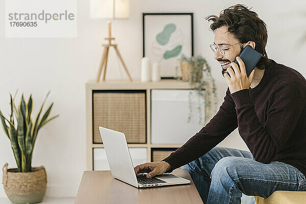 Glücklicher Mann mit Laptop  der zu Hause mit dem Handy telefoniert