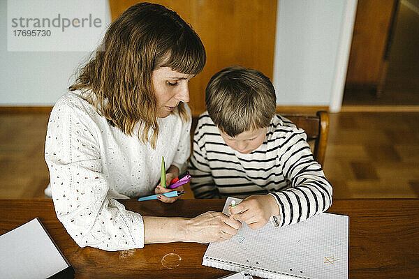 Blickwinkel der Mutter  die ihrem Sohn mit Down-Syndrom das Zeichnen in einem Buch am Tisch beibringt