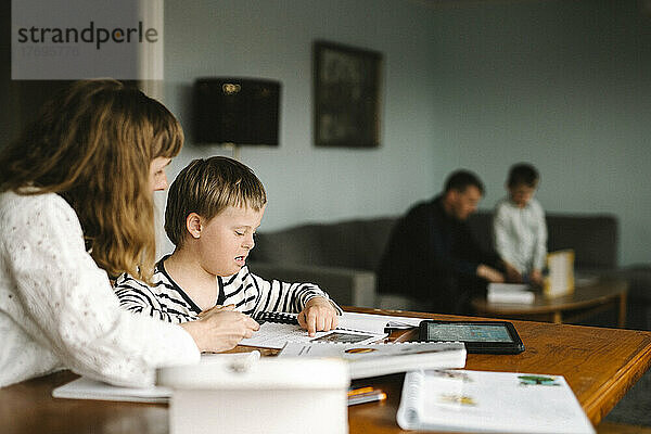 Junge mit Down-Syndrom  der von seiner Mutter ein Buch liest  während er am Tisch Hausaufgaben macht