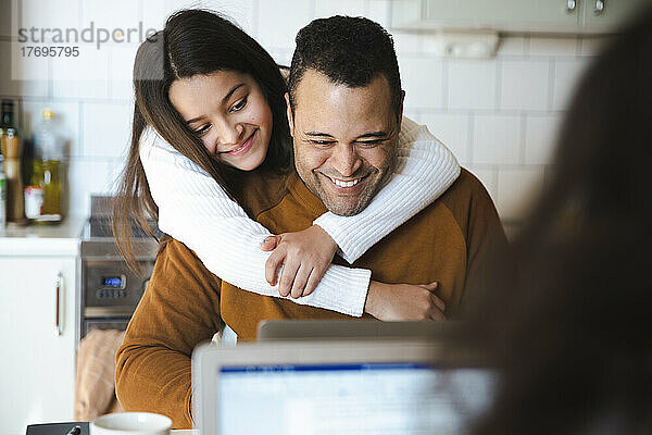 Lächelndes Mädchen umarmt Vater  der zu Hause am Laptop arbeitet