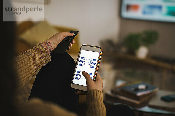 Ausgeschnittenes Bild einer Frau  die eine App auf ihrem Smartphone benutzt  während sie zu Hause fernsieht