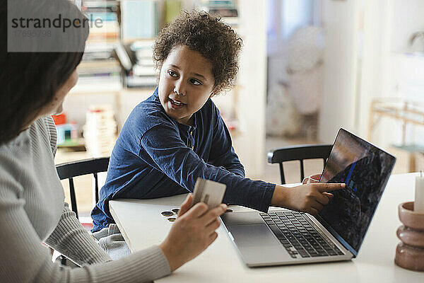 Der Sohn zeigt auf den Laptop-Bildschirm  während er seine Mutter mit der Kreditkarte ansieht
