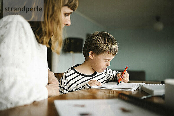 Junge mit Down-Syndrom schreibt in ein Buch  während er mit seiner Mutter am Tisch sitzt