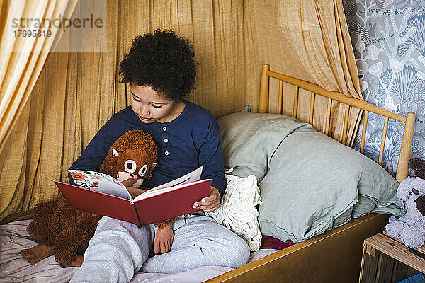 Junge mit Stofftier  der zu Hause auf dem Bett sitzend ein Buch liest