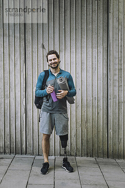 Ganzkörperporträt eines Mannes mit Behinderung  der eine Trainingsmatte und eine Wasserflasche vor einer Wand trägt