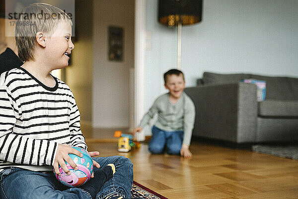 Glücklicher Junge mit Down-Syndrom mit Ball auf dem Boden sitzend im Wohnzimmer