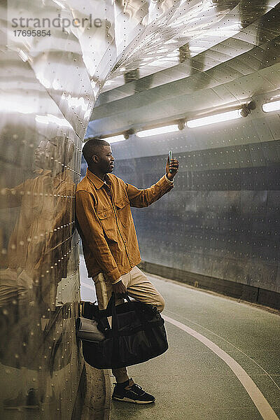 Junger Mann in voller Länge  der eine Tasche trägt  während er ein Selfie mit seinem Mobiltelefon in einem U-Bahn-Tunnel macht