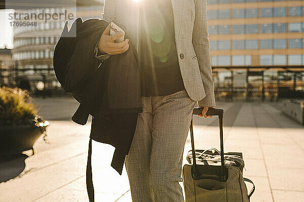 Midsection von Geschäftsfrau mit Smartphone ziehen Gepäck beim Gehen auf sonnigen Tag