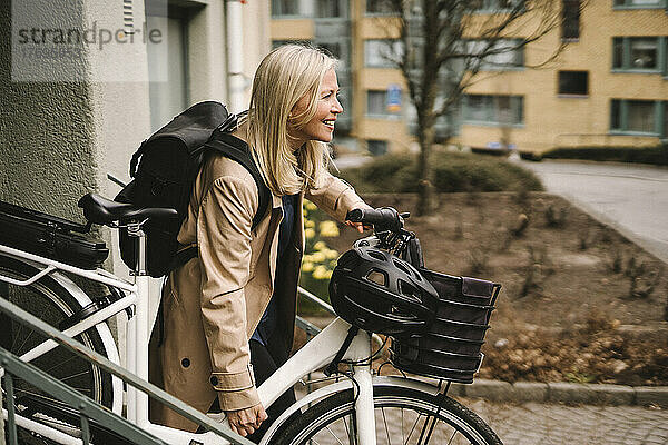 Seitenansicht einer lächelnden Geschäftsfrau mit Rucksack  die ein Elektrofahrrad hält