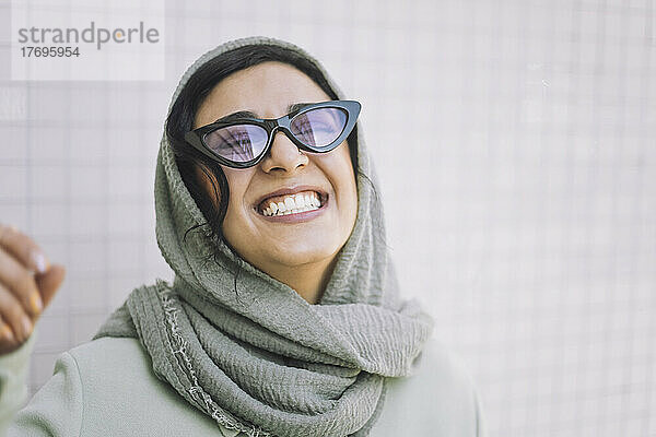 Fröhliche Frau mit Kopftuch und Sonnenbrille an der Wand