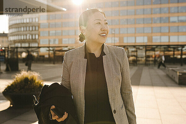 Lächelnde Geschäftsfrau  die mit Mantel an einem sonnigen Tag steht und wegschaut