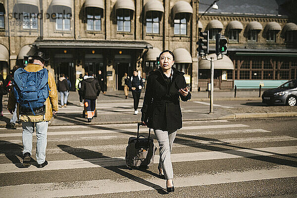 Geschäftsfrau zieht Gepäck beim Überqueren der Straße in der Stadt an einem sonnigen Tag