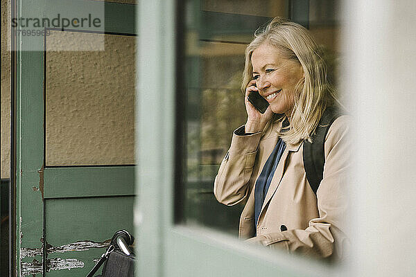 Lächelnde Geschäftsfrau  die mit einem Smartphone spricht  gesehen durch ein Glasfenster